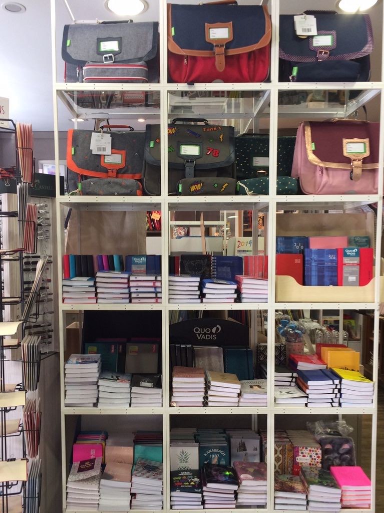 Une étagère blanche de la boutique: des cartables en haut, des agendas et des carnets empilés en dessous