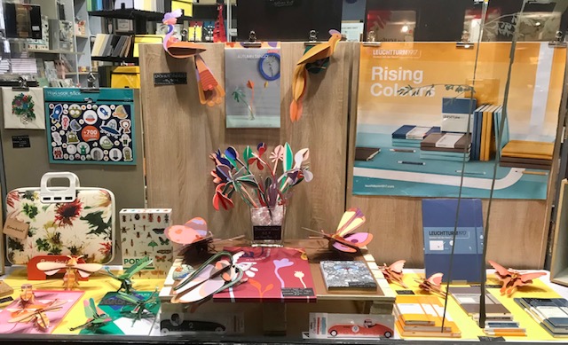 Une vitrine de Lancryer avec des fleurs et des oiseaux en carton coloré, une pochette fleurie et des carnets